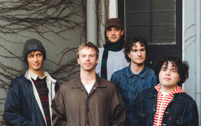 The band is made up of five men. They are stood in front of the door to a house an looknig at the camera.