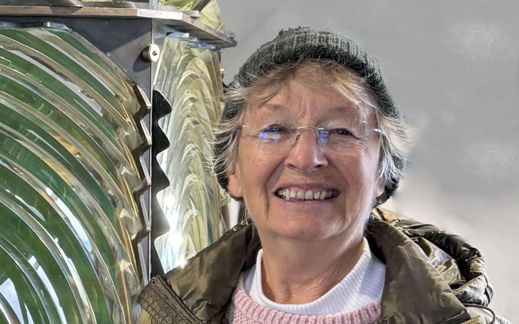 This is a head and shoulders shot of Anne Rimmer. She is weating a green puffer jacket and clear, frameless glasses. Anne is standing next to an old lightouse lens.