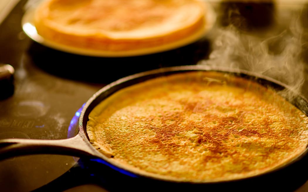 Image of a crepe cooking in a cast iron crepe pan with a stack of crepes in the background