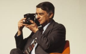 Edwin Land sits in an orange chair wearing a brown suit and holding a polaroid camera up to his face.