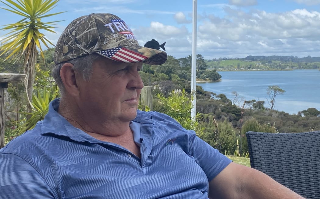 The Mayor of Kaipara sitting on his deck looking out at the mangroves with a displeased expression and a camouflage Trump hat.