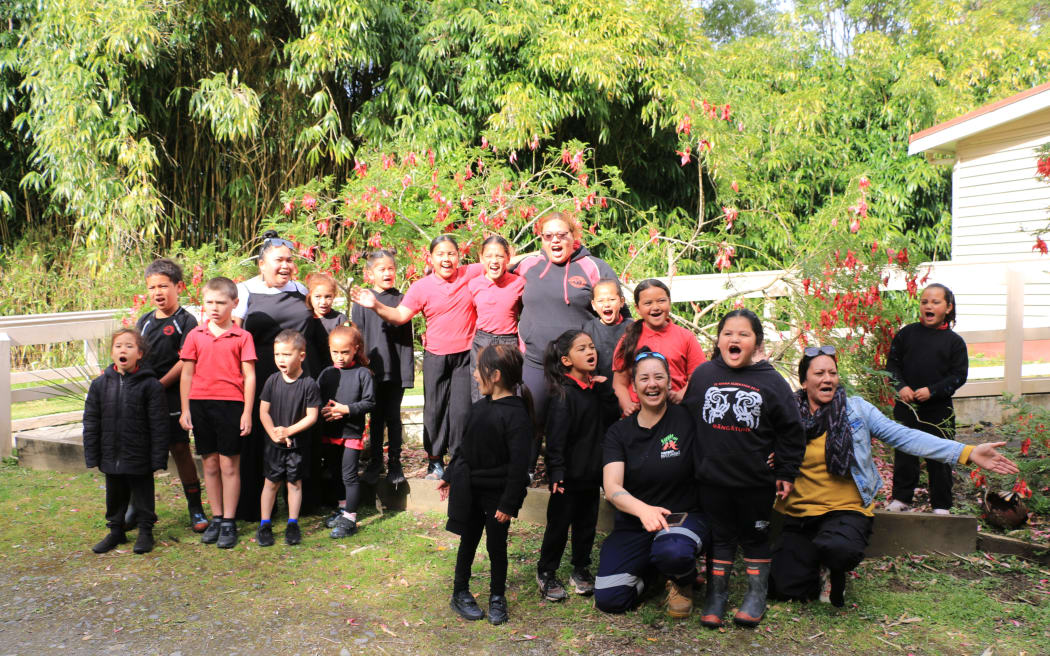 A group of about 15 children in black-and-red uniforms with their teachers gather for a group photo in front of a wooden fence and green bushes with bright red flowers.
