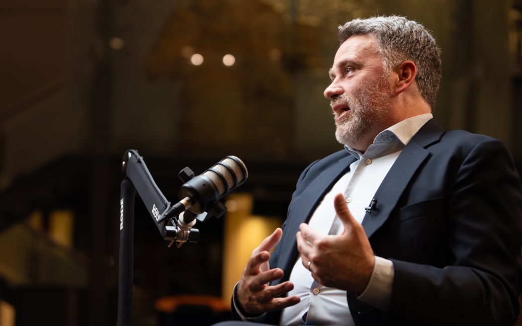 A man sits behind a microphone on a stand, he is explaining something and gesturing with his hands.