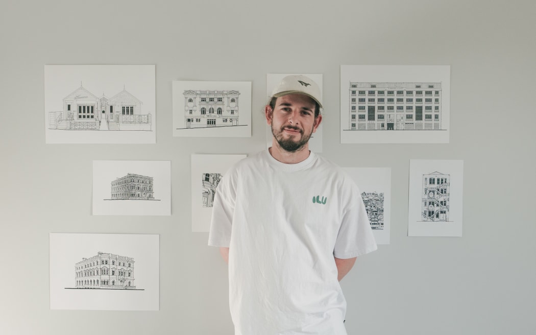 Josh stands in front of a white wall with prints of his illustrations tacked on to it.