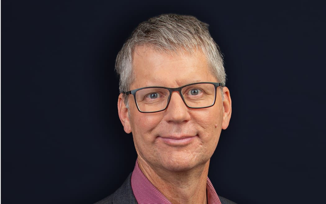 Bryan Crump looks down the camera with an open expression, wearing glasses and a dark pink shirt and jacket