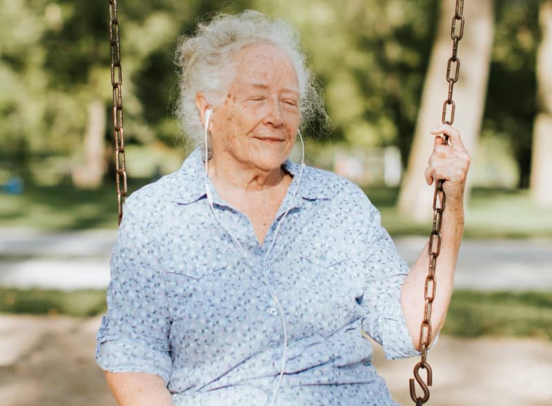 A senior woman sits on a swing smiling as she listens to music through wired headphones.