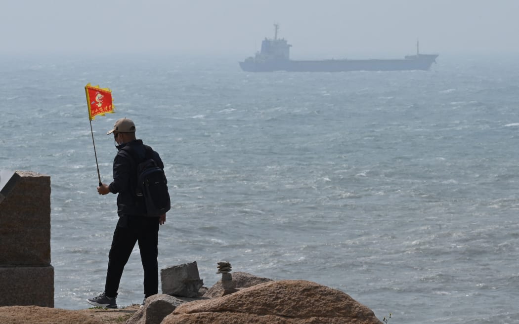 A man walks with a flag as a ship passes by behind him on Pingtan island, the closest point to Taiwan, in China’s southeast Fujian province on April 8, 2023. - China launched military drills around Taiwan on April 8, in what it called a "stern warning" to the self-ruled island's government following a meeting between its president and the US House speaker. (Photo by GREG BAKER / AFP)