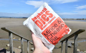 A hand holding up a Maketu Pies mince and cheese pie in its wrapping in front of a beach.