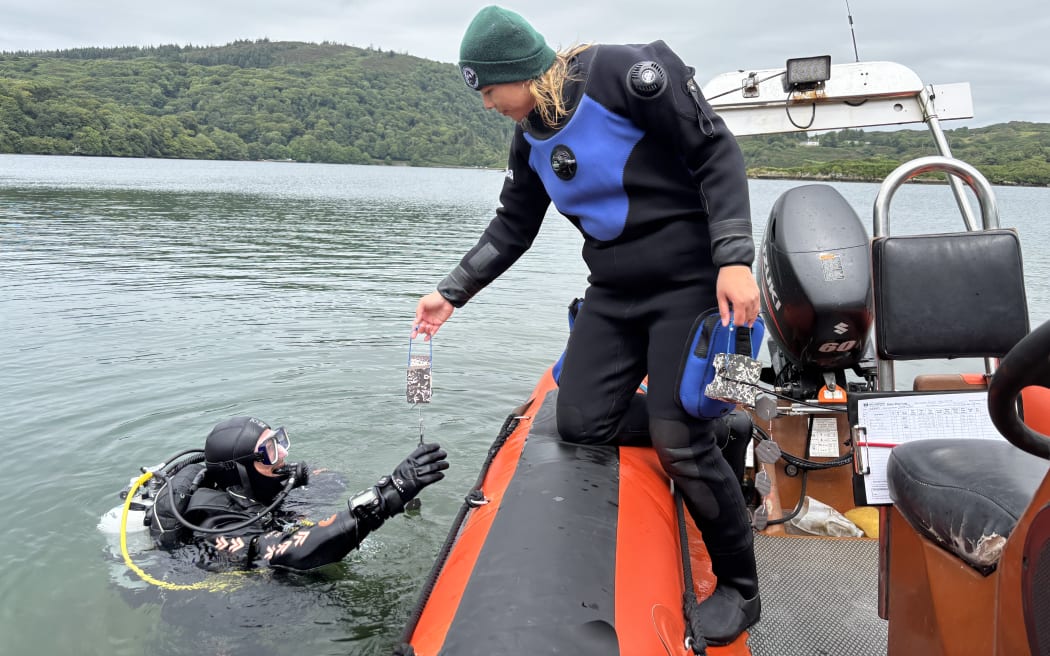 A diver in the water (James) receives a weighted set of tiles from another diver on a boat (Kea). The background features green hills and an overcast sky.
