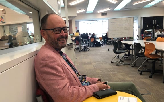 Maurie Abraham smiles at the camera. He is sitting side on from the camera in a chair, with a classroom behind him.