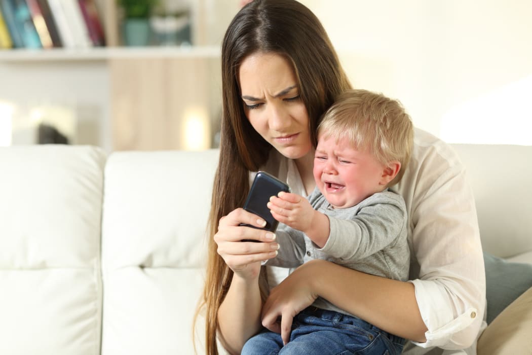 A photo of a baby having a tantrum and fighting with his mother for a smart phone sitting on a couch in the living room at home