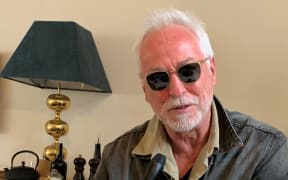 Eddie Raynor holding a microphone at a kitchen table, a lamp and teapot in the background.