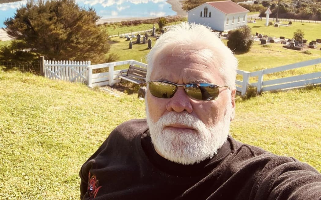 Phil Paikea wears sunglasses and stands in front of a house, a lake, mountains and a blue sky