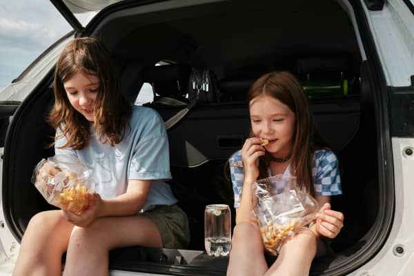 Two kids sit in the open boot of a car, eating snacks from a ziplock bag.