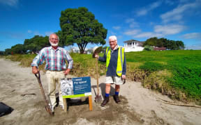 Two men stand with shovels on a beach next to a small wooden sign. The sign reads "I NEED MY SPACE!" next to a photo of a dotterel. It is a fine day, with blue skies.