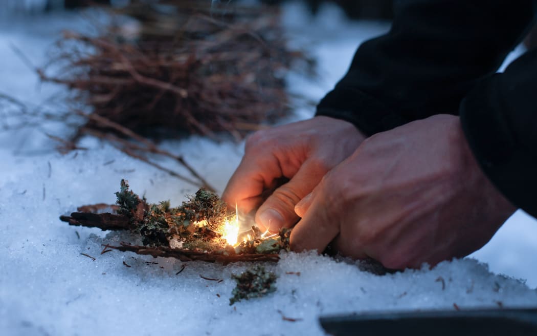 Man lighting a fire in a dark winter forest, preparing for an overnight sleep in nature, warming himself with DIY fire. Adventure, scouting, survival concept.