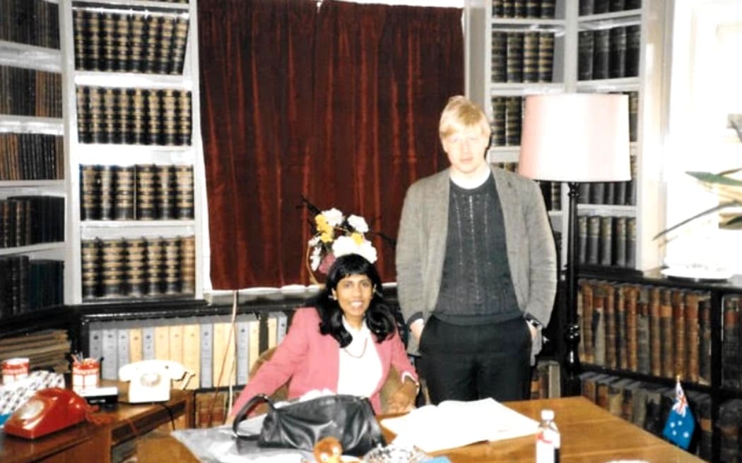 Jeya and Boris are inside an office. The walls are lined with bookshelves holding heavy volumes. Jeya sits at a large wooden desk, wearing a pink blazer and smiling. Boris stands beside her, wearing a grey suit jacket, with his hands in his pockets.