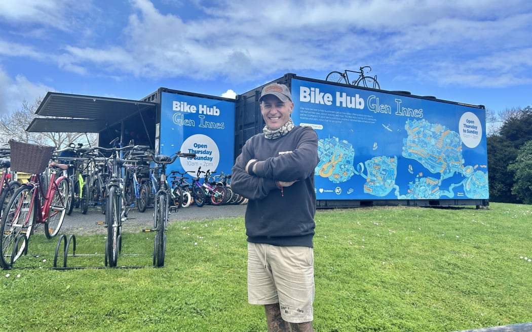 Portrait of a 47 year old man standing on the grass outside a bike hub, with second hand bikes in the background.