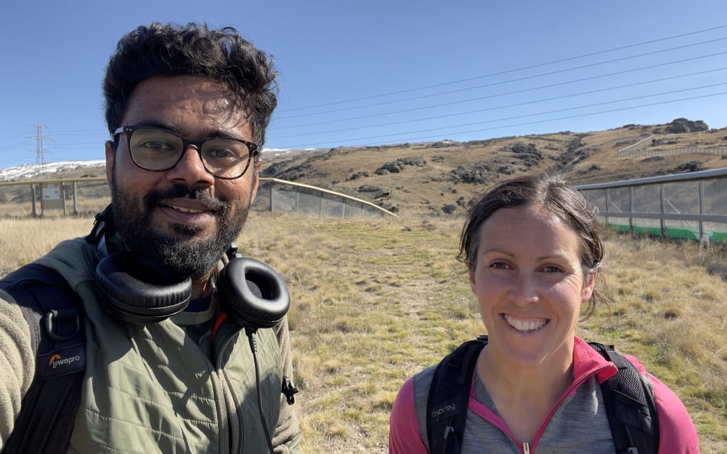 A man with headphones around his neck takes a selfie with a woman smiling and wearing a backpack. They are standing in a dry grassy field with a high, mesh predator proof fence in the background below a rolling brown hill beneath a blue sky.