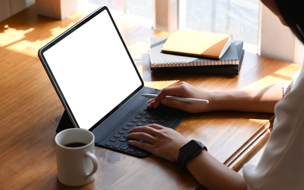 Cropped image of young beautiful woman working as writer typing on computer tablet with white blank screen while sitting at the wooden working table with sunlight through windows as background.
