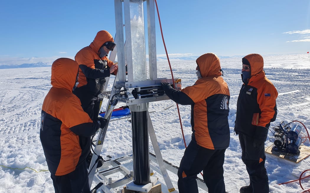 Four scientists in Antarctica, wearing orange and black jackets. They are standing around a piece of silver equipment.
