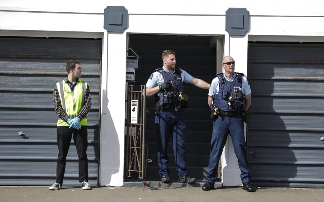 Two uniformed police and a person in a high vest vest with blue gloves stand outside a doorway.