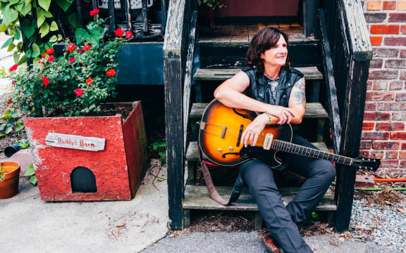 Singer-songwriter Amy Ray sits on outside steps with her guitar; red flowers in a planter box nearby.