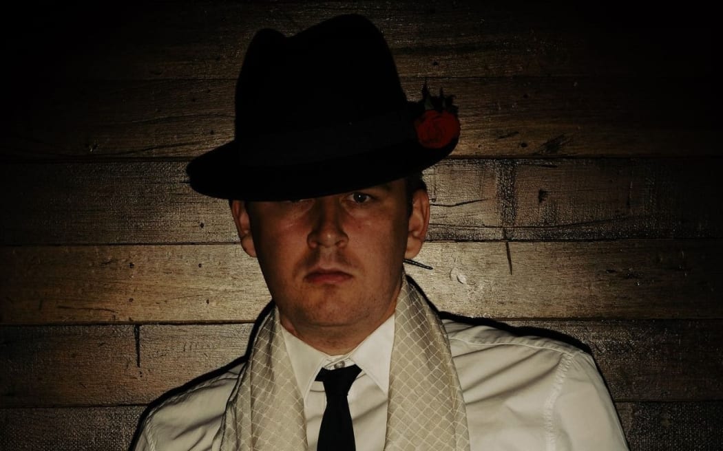 Richard III (Chris Raven) faces the camera in a white shirt and vintage black hat.