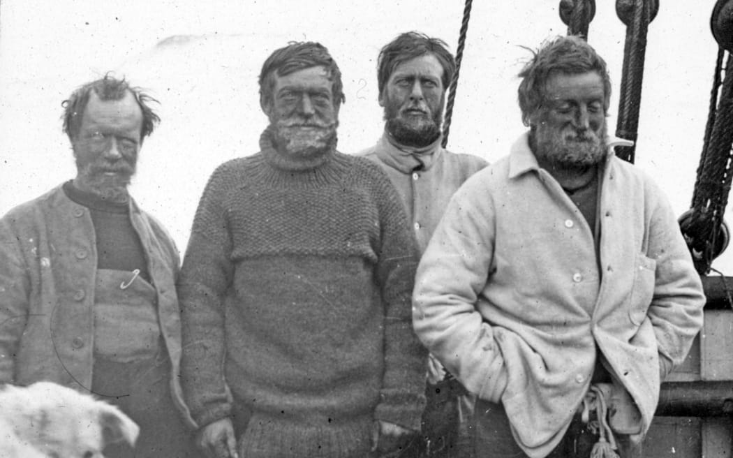 A historic photo showing four men from the Nimrod expedition: Frank Wild, Ernest Shackleton, Eric Marshall, and Jameson Adams. The men are stood on a ship, and are all wearing thick woolen jumpers and jackets.