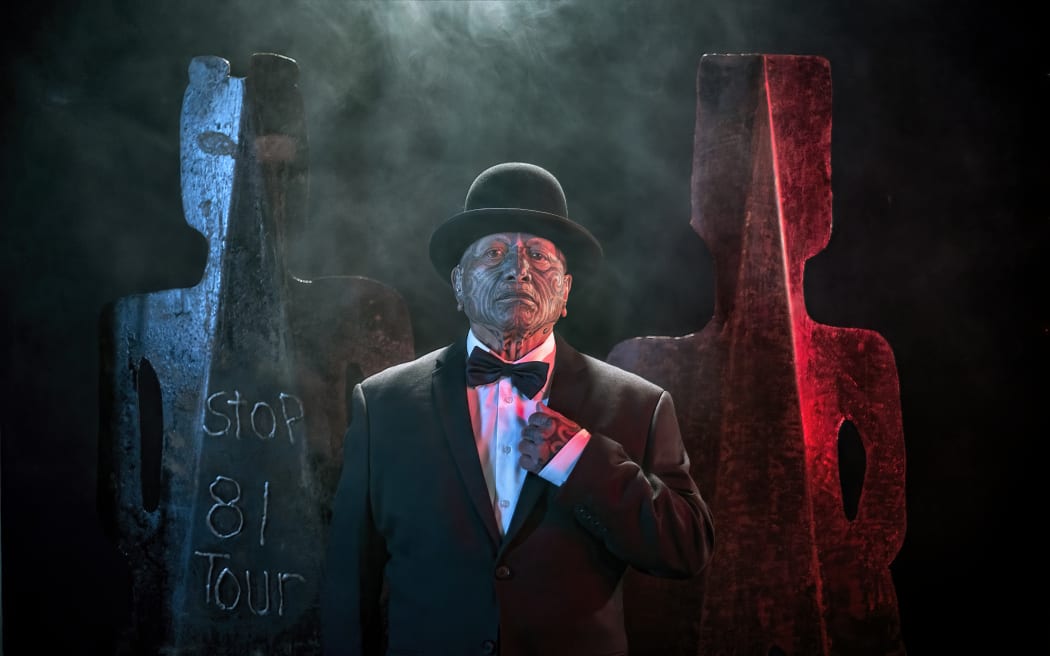 Image from Tame Iti: I Will Not Speak Māori Exhibition.  Artist wearing a suit, bowtie and bowler hat stands in front of metal sculptures in a stylised human form.