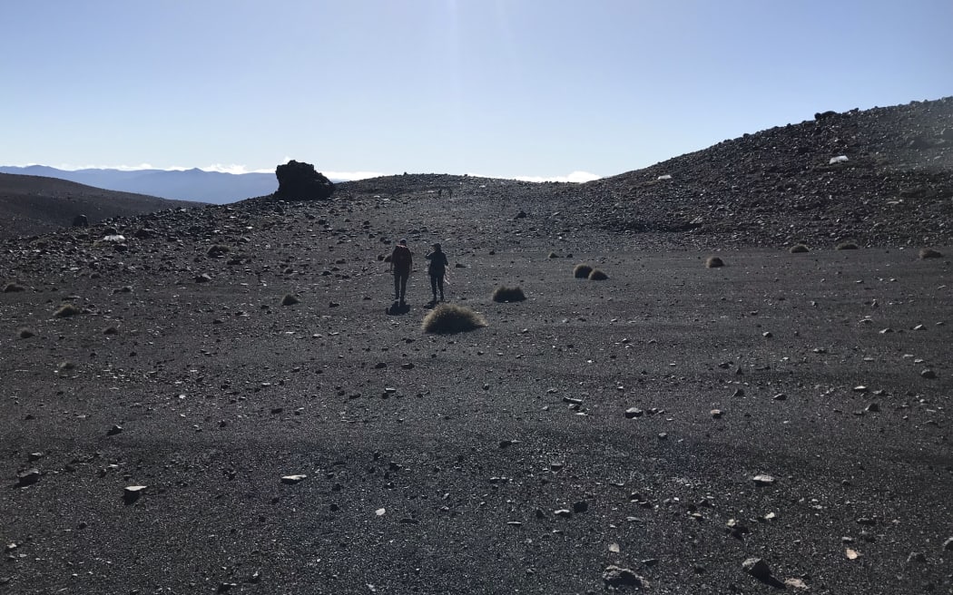 Two people holding equipment stand in the mid ground. The scenery is dark gravel and rocks, it looks very much like a moonscape, with a smattering of plants. There's a clear blue sky in the background.