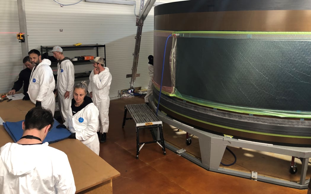 A group of workers in white jumpsuits are busy at a table. Behind them is an enormous metal cylinder.