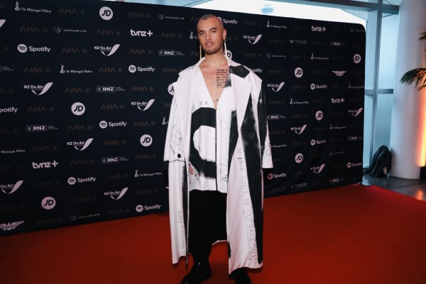 Stan Walker, a shaven-headed man, wears a large black and white coat while standing on a red carpet.