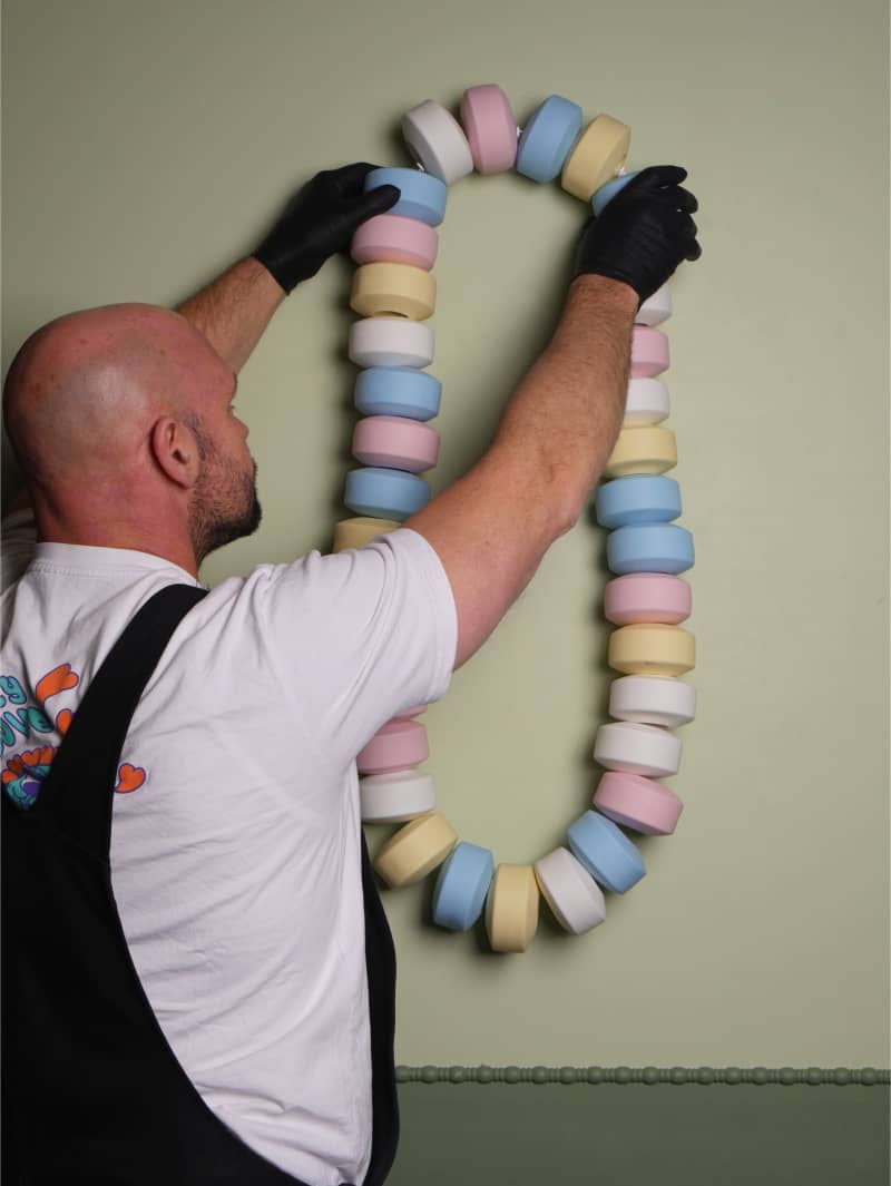 A bald man in a white t-shirt arranges a colourful artwrk on the wall.