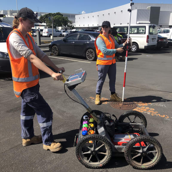 Two women, one with a buggy-like piece of equipment (a ground penetrating radar unit) and the other holding a stick-like piece of equipment (a GPS locator).