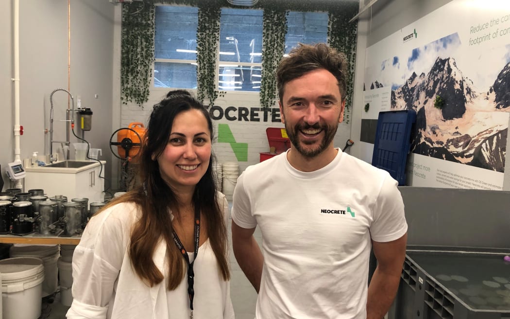 Zarina and Matt smile at the camera. They are standing inside an office room decorated with colourful posters and lab equipment.