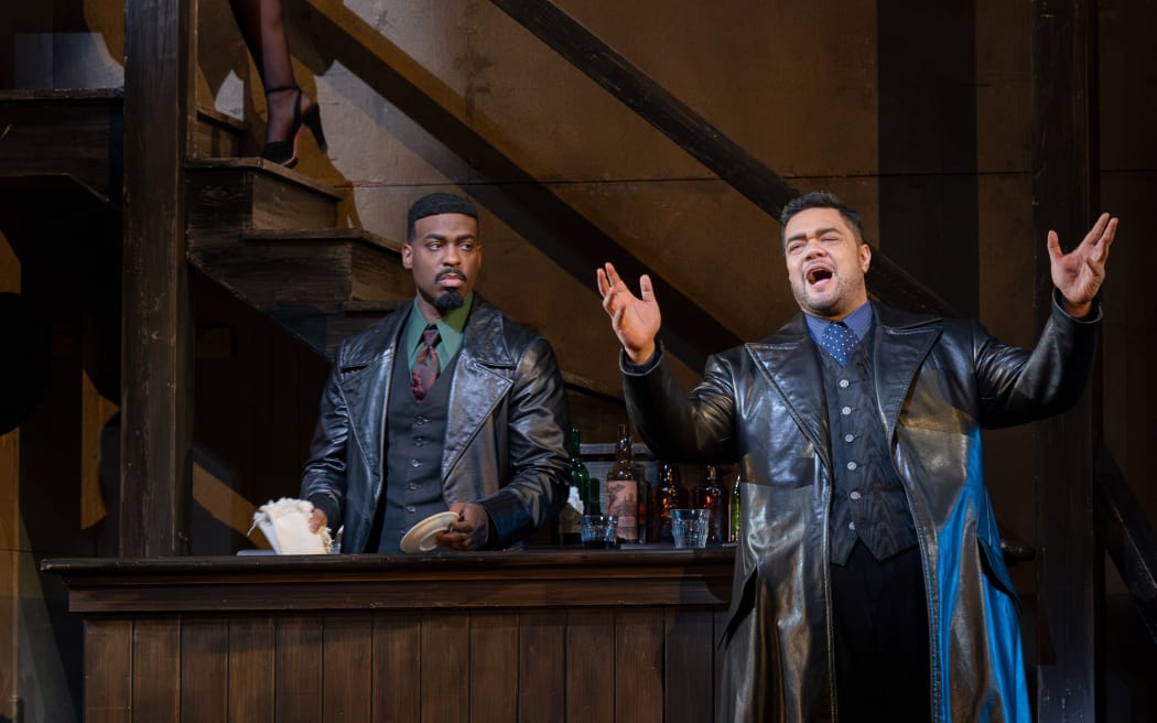 Pene stands on stage singing. He wears a large leather coat over a suit. Other actors glare at his character as he sings grandiosely.