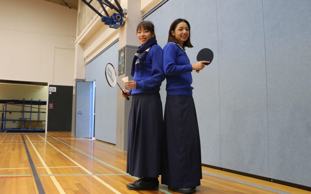 Two young women stand back to back insports hall with bat and racquet