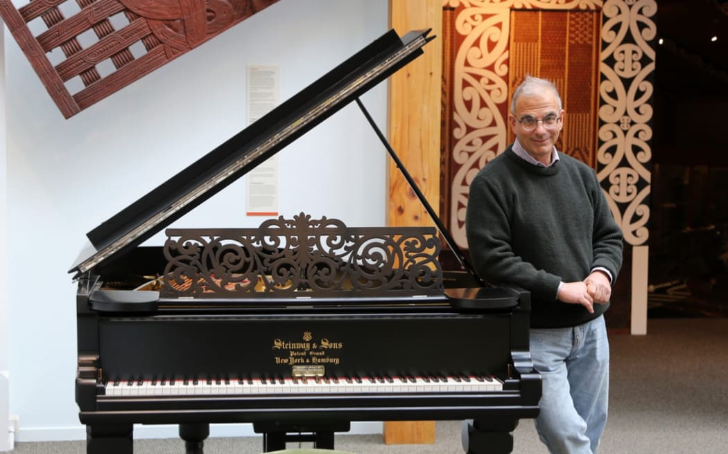 A man stands next to a old style piano. He has short grey hair and is wearing glasses with clear rims, he is dressed in a green sweater and light blue jeans.