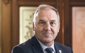 Andrew smiles at the camera. He wears a navy suit with a navy tie. He is sitting in a room with wood pannelling.