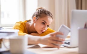 A bored-looking girl looks at her phone as she leans on desk with laptop open.