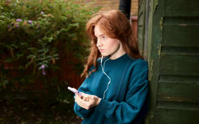 A worried-looking redhead teenager holds her phone and listens to something on wired headphones