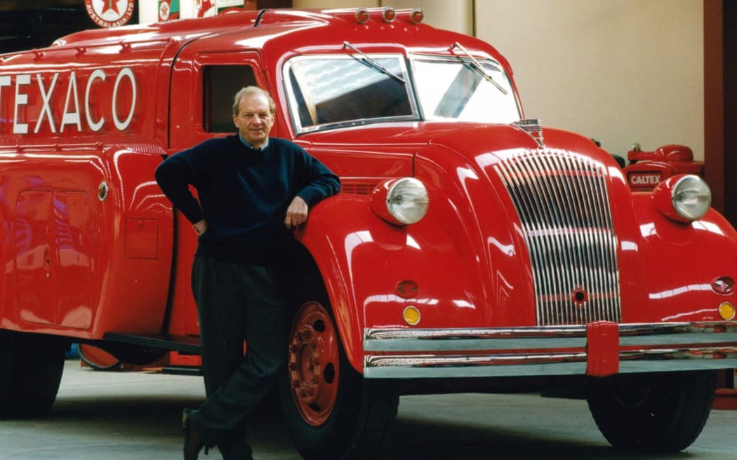 Bill stands in front of a large red car, resting his elbow on the hood, smiling.