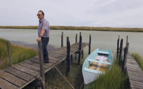 Man stands on small wooden jetty on edge of murky river by small wooden boat