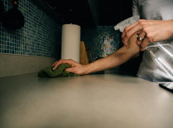 Person wiping down a kitchen bench with a cloth and spray in other hand.