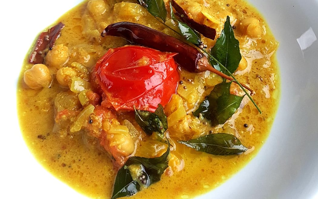 A close-up image of coconut chickpea curry on a shallow white bowl, decorated with curry leaves and a tomato.
