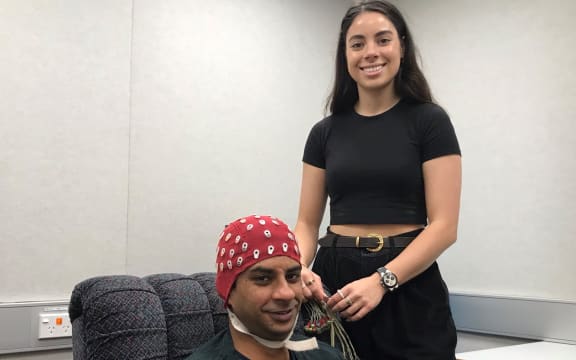 Dunja is smiling, wearing a black crop top and jeans, holding a bundle of wires. Beside her Amit sits in a comfy chair, with the red EEG cap on his head, you can see the labelled holes where the wires will be attached.