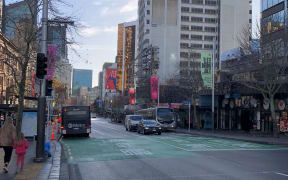 Queen Street on a quiet weekday afternoon. The sky is pale blue, the streets are in shadow. A few buses and cars busy the roads. A woman and her daughter walk along the sidewalk on the left.