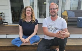 Two friends, a woman and a man are sitting side by side on the outside steps of a deck, smiling at the camera. They are barefoot, casually dressed and look relaxed.