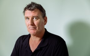 Jeremy Buxton sits for a photograph, his shadow being cast on the wall behind him.